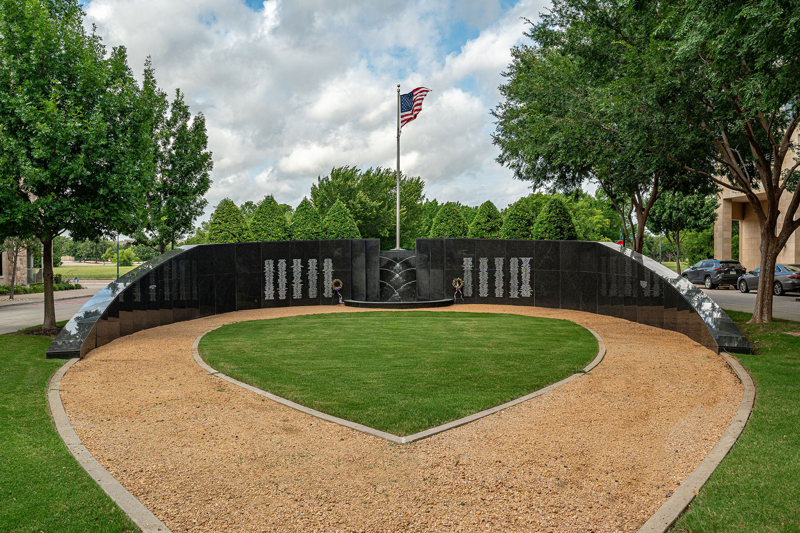 A curved black granite memorial wall engraved with names stands on a grassy area; an American flag on a pole is centered behind the wall, surrounded by trees and parked cars.