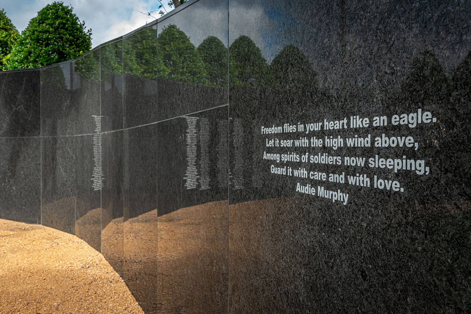 A curved black granite memorial wall with engraved text, including a quote by Audie Murphy, stands beside a gravel path and green trees.