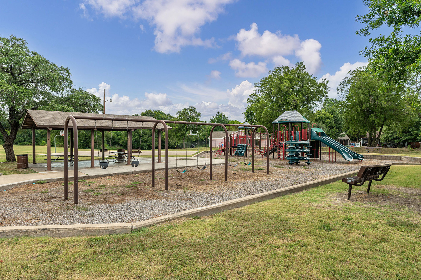 Outdoor playground with empty swings, slides, climbing structures, and a covered picnic area, surrounded by grass and trees on a sunny day.