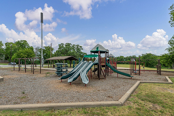 Empty playground with multiple slides, climbing structures, monkey bars, and a covered seating area, set on gravel with grassy areas and trees in the background under a partly cloudy sky.