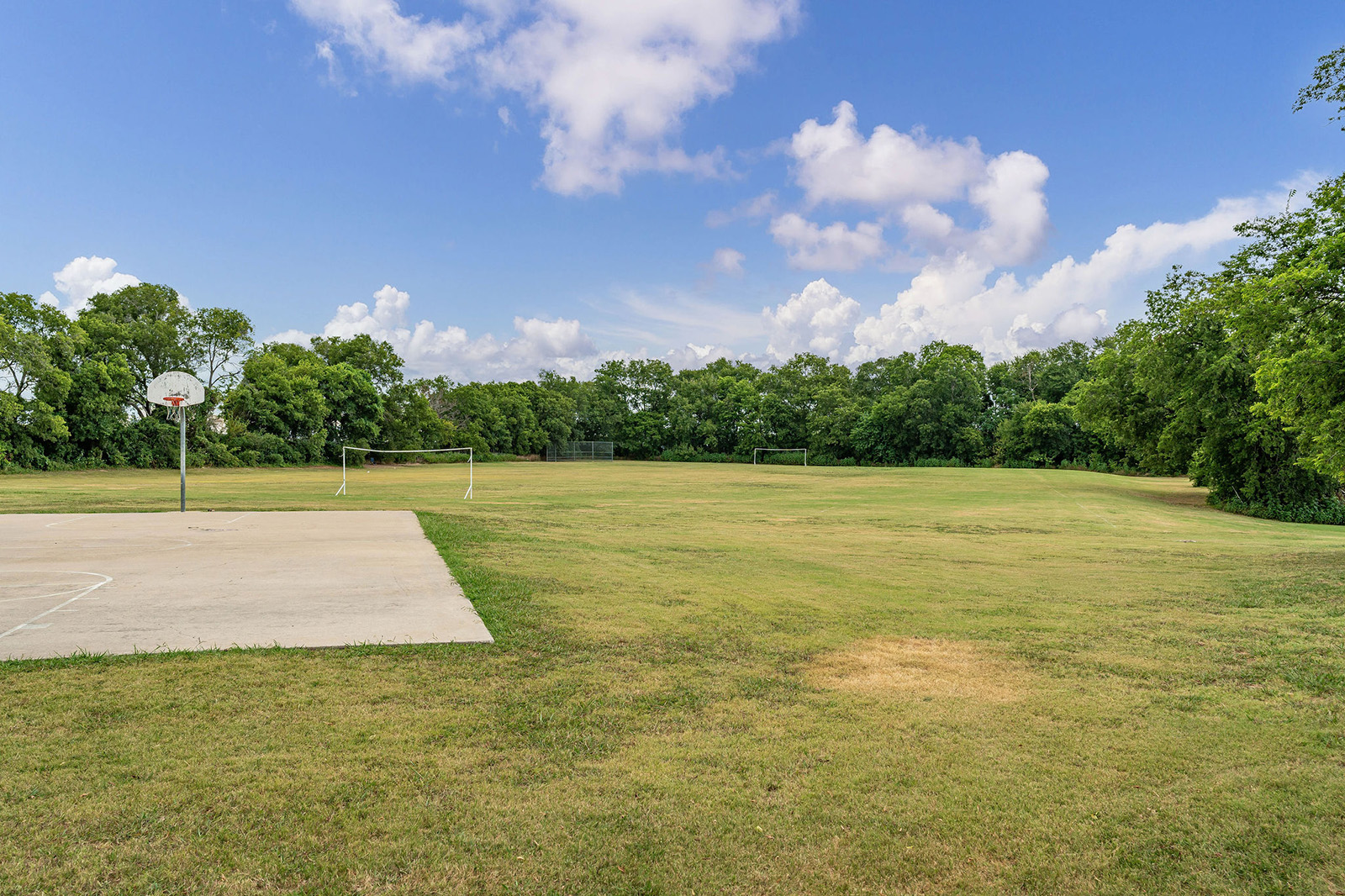Outdoor sports field with a grassy area, two soccer goals, and a concrete basketball court, surrounded by trees under a partly cloudy sky.