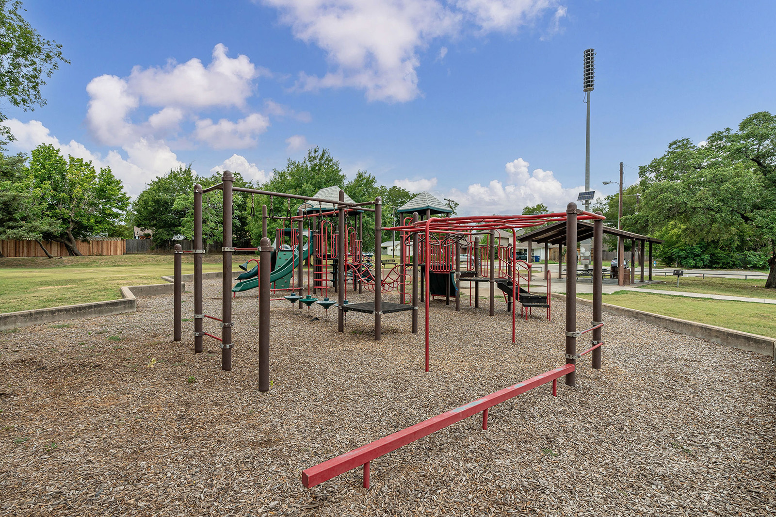 Outdoor playground with climbing structures, slides, and monkey bars on woodchip ground, surrounded by trees and grassy areas under a partly cloudy sky.