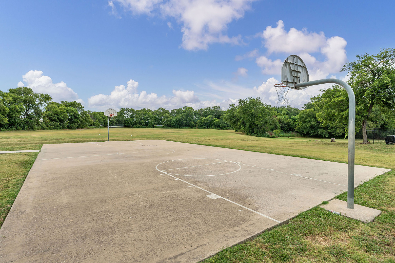 Outdoor basketball court with two hoops, surrounded by grass and trees under a partly cloudy sky.