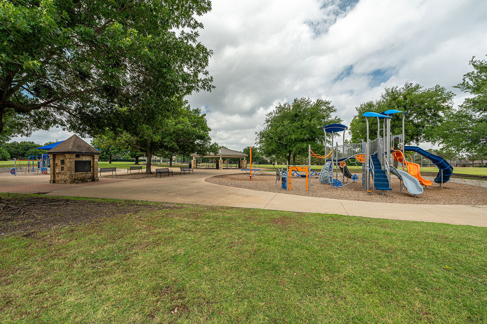 Outdoor playground with slides and climbing structures on mulch, surrounded by trees, a gazebo, benches, and a paved walkway on a cloudy day.