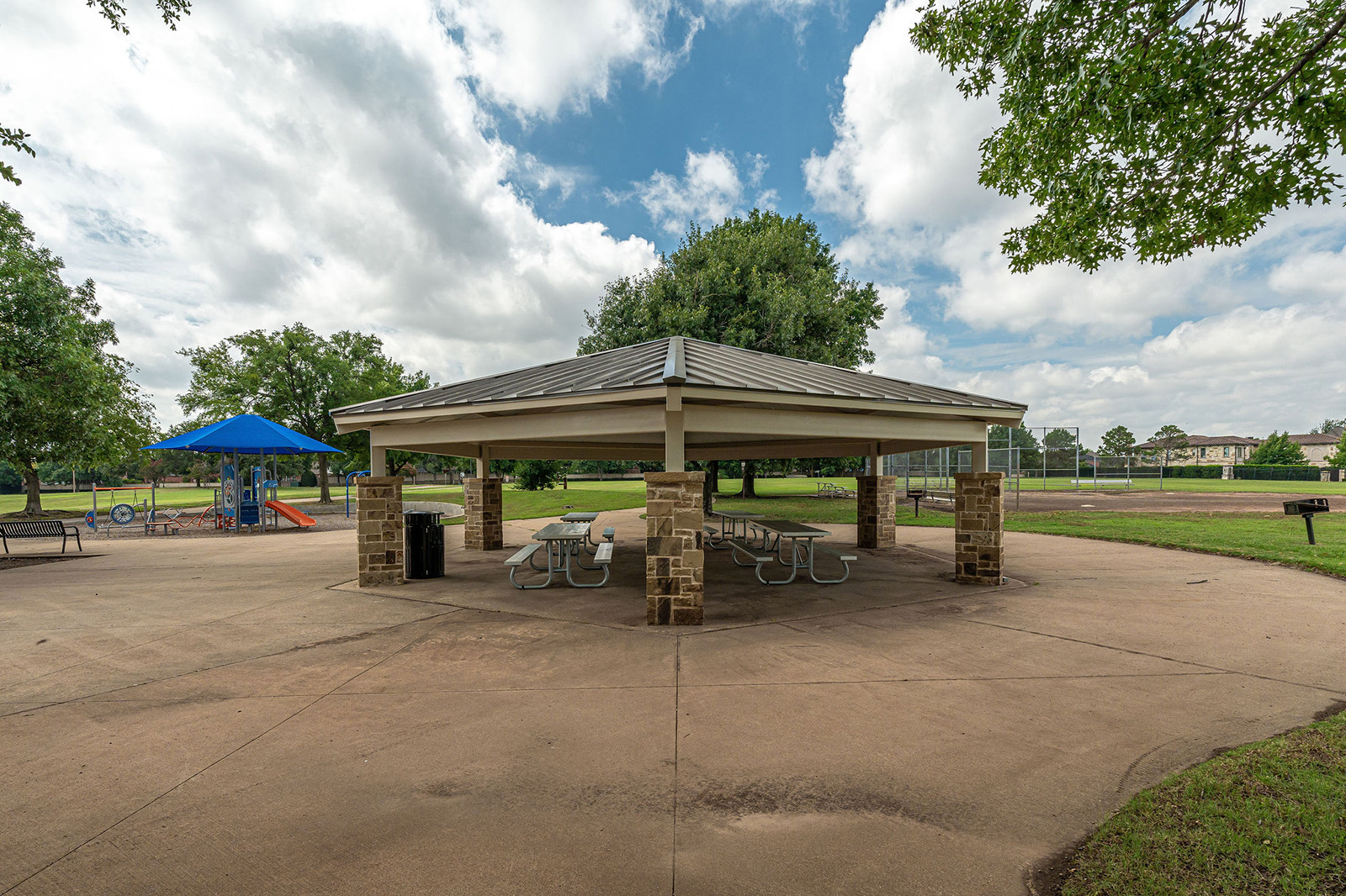 A covered picnic pavilion with tables and trash cans is in the center of a park. A playground and trees are visible in the background under a partly cloudy sky.