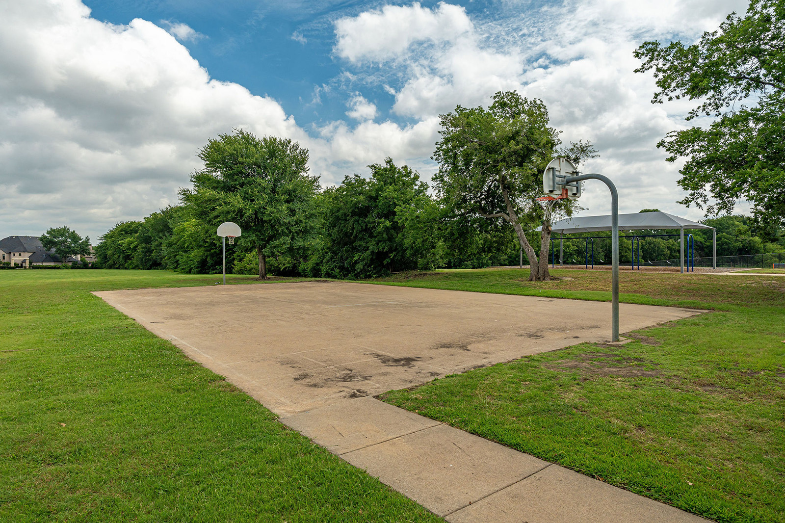 Outdoor basketball court with two hoops, surrounded by grass and trees, under a partly cloudy sky. A covered pavilion is visible in the background.