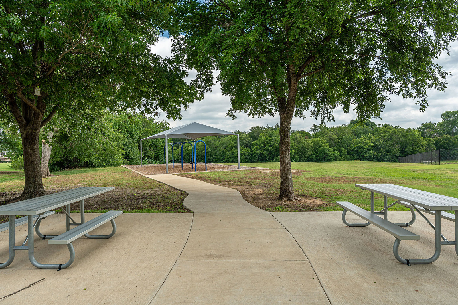 A concrete path leads to a covered playground structure in a grassy park, with picnic tables and trees in the foreground.