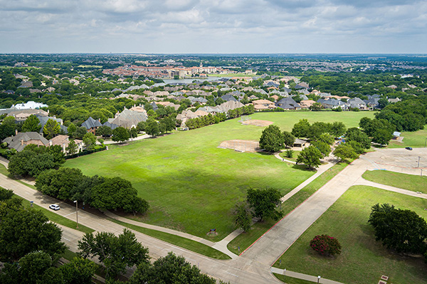 Aerial view of a suburban neighborhood with houses surrounding a large green park, intersecting roads, and scattered trees under a partly cloudy sky.