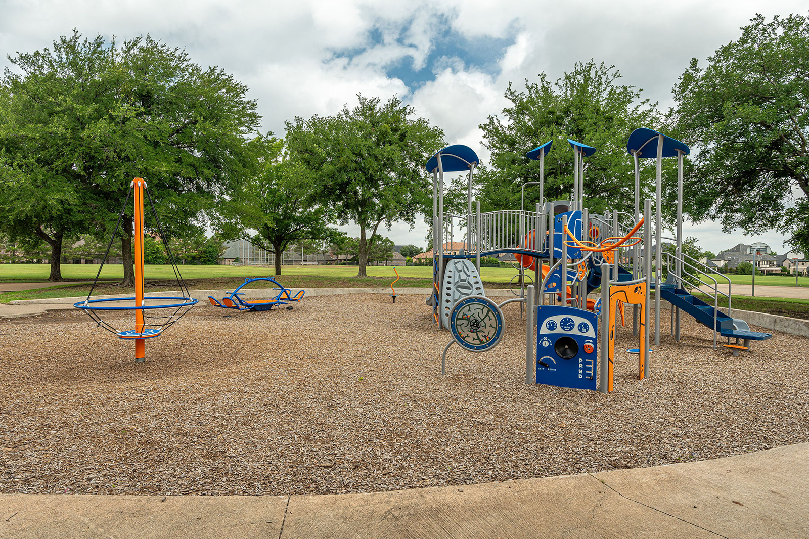 Outdoor playground with blue and orange equipment, including slides, climbing structures, and swings, set on wood chips with trees and grass in the background.