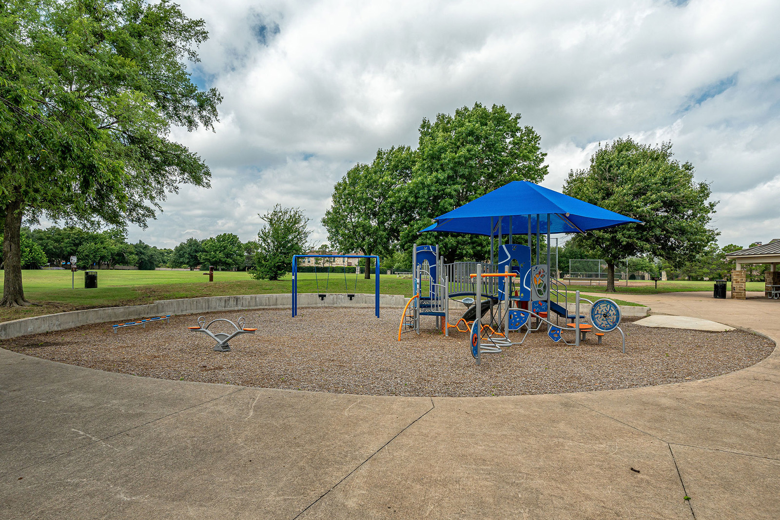 A playground with blue equipment and a shade canopy is set on a gravel surface, surrounded by trees and grassy fields under a partly cloudy sky.