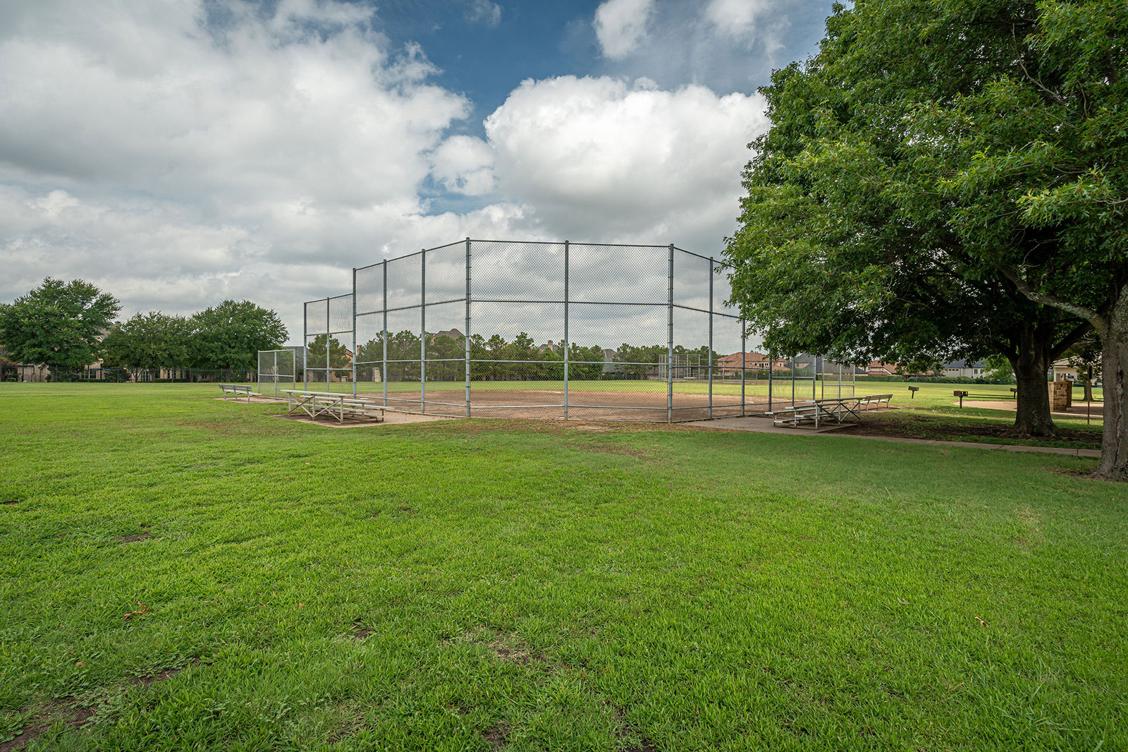 An empty baseball field with a backstop, metal bleachers, and green grass, bordered by trees under a partly cloudy sky.