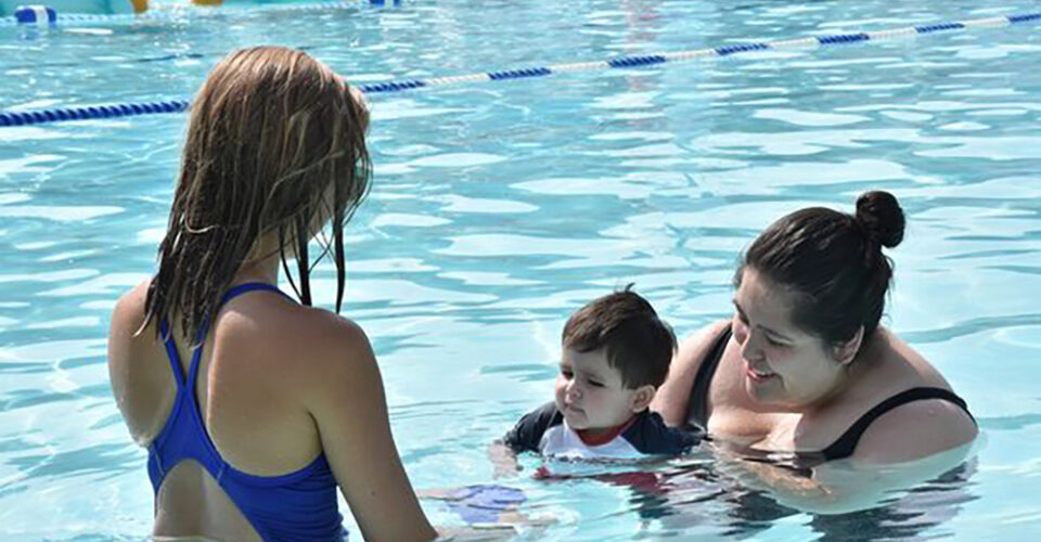 A woman holds a baby in a swimming pool while another woman in a blue swimsuit stands nearby.