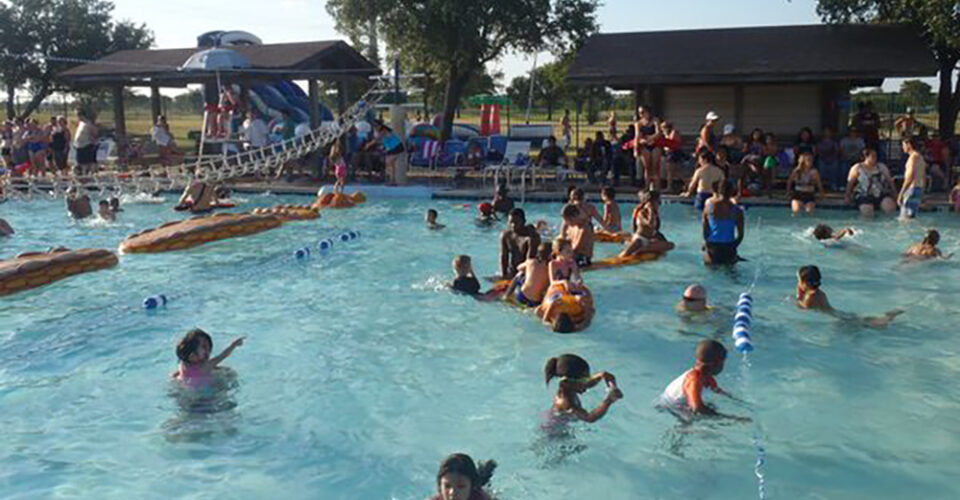 Dozens of people, including children and adults, swim and play in a crowded outdoor pool on a sunny day, with pool floats and shaded pavilions nearby.