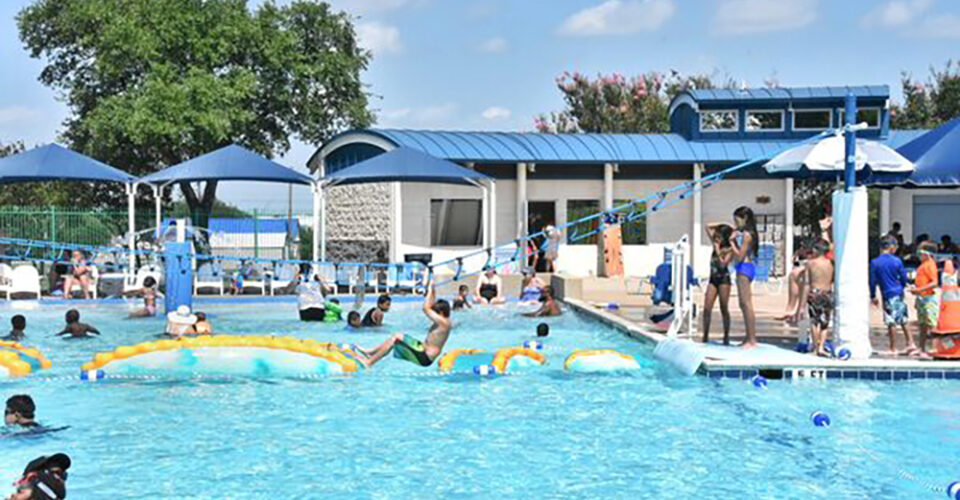 People swim and play on inflatable obstacles in an outdoor public pool, with shaded areas, trees, and a blue sky in the background.