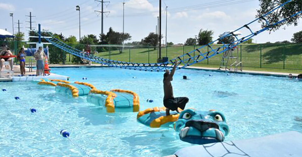 A person hangs from a rope above a swimming pool, aiming to land on a floating dragon-shaped obstacle. Other people are in and around the pool.