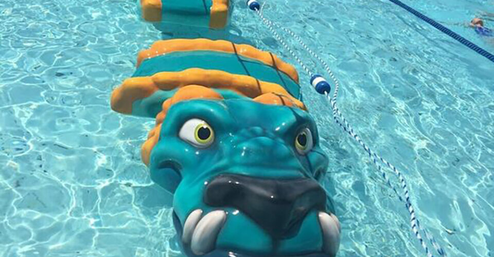 A blue and yellow inflatable dragon float is placed in a swimming pool, positioned parallel to a swim lane divider.