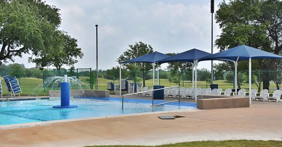 Outdoor swimming pool with a shallow water area, blue sun shades, white lounge chairs, and green trees in the background on a cloudy day.