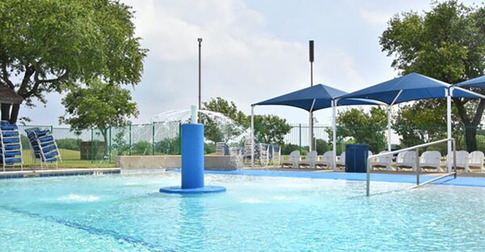 Outdoor swimming pool with lounge chairs, blue shade canopies, and trees surrounding the area on a partly cloudy day.