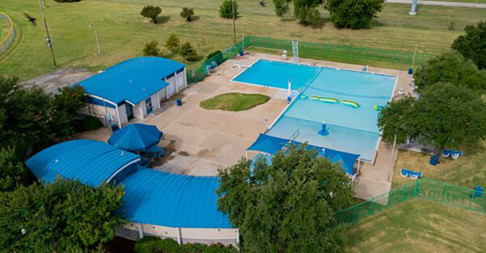 Aerial view of an outdoor public swimming pool complex with blue roofs, surrounded by grassy fields and trees.
