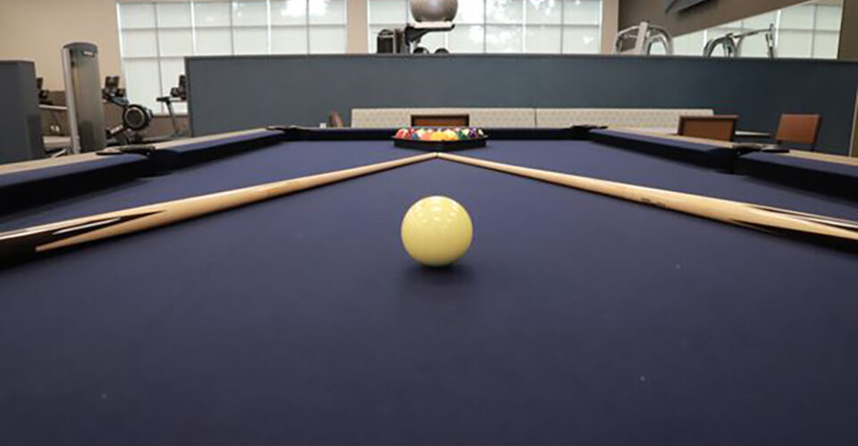 A pool table with a cue ball in the foreground, four pool cues arranged diagonally, and racked balls in the background in a gym setting.