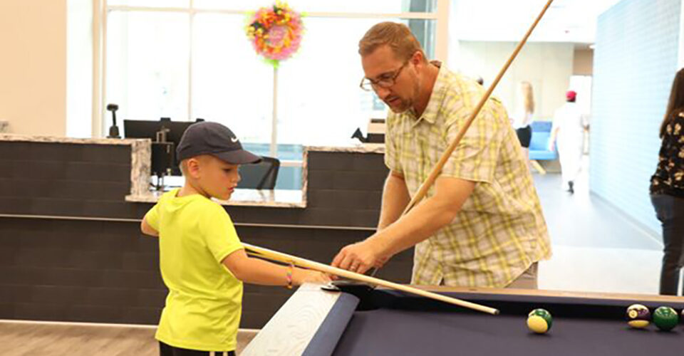 An adult teaches a young boy in a neon shirt and cap how to aim a pool cue at a billiards table in a well-lit indoor space.