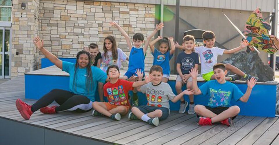 A group of children and one adult sit and kneel on a wooden platform outside, smiling and raising their hands in the air.