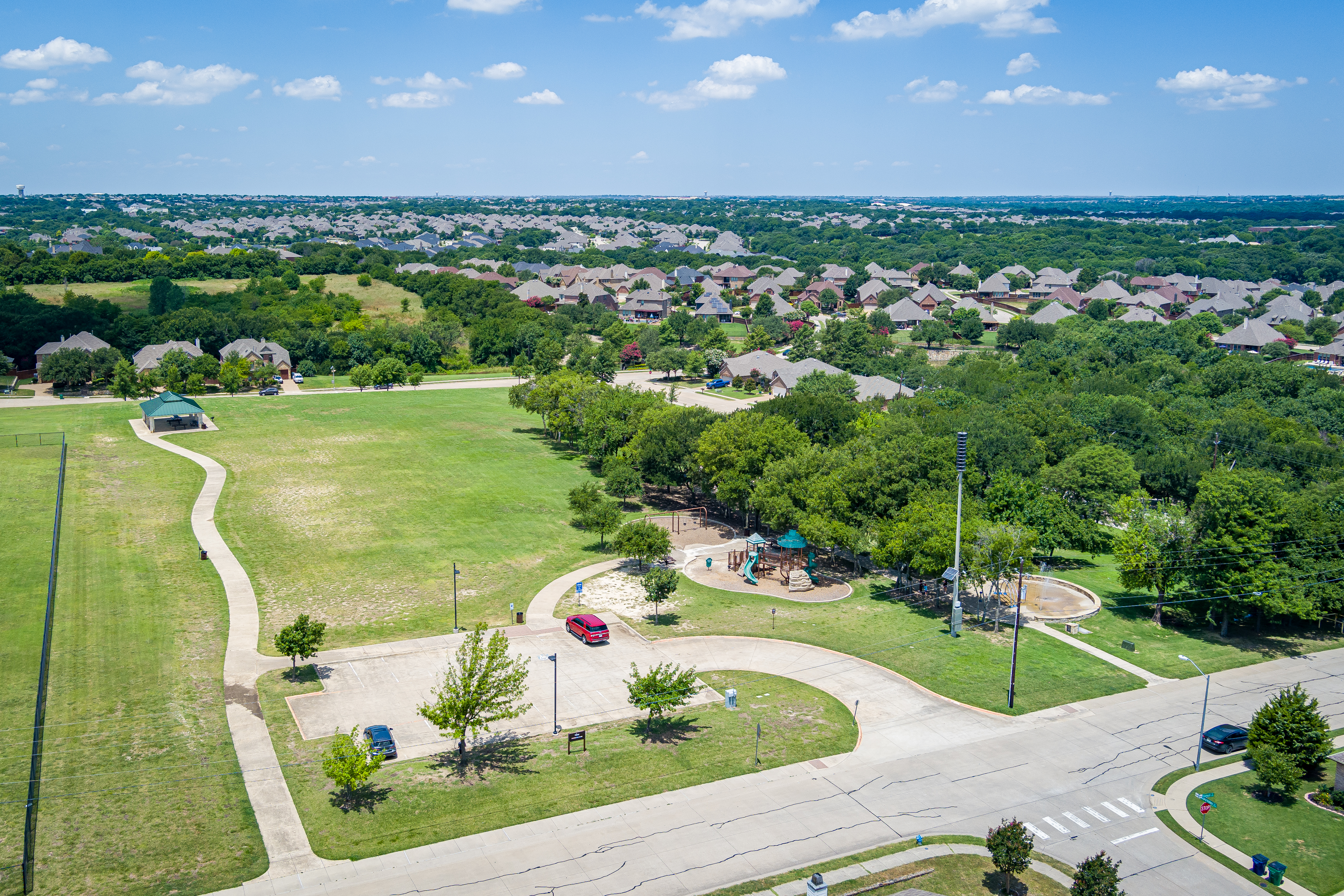 Aerial view of a suburban park with green fields, curving pathways, playgrounds, trees, and nearby houses under a blue sky with scattered clouds.