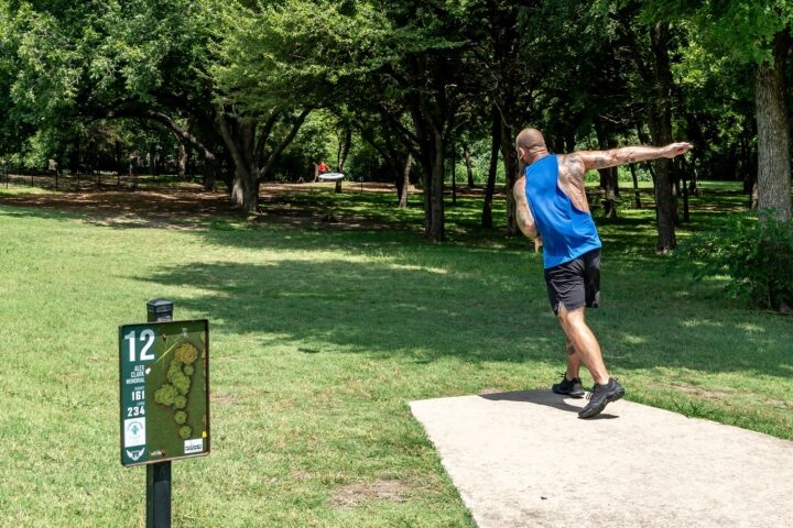 Alex Clark Disc Golf with a tee box sign number 12 and a guy throwing disc on concrete pad.